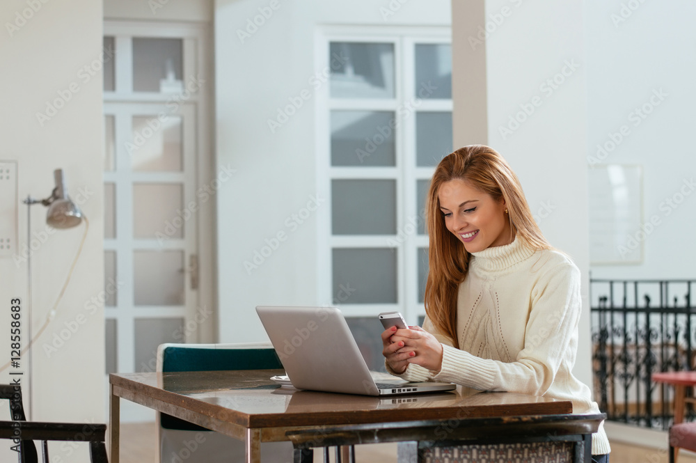 Attractive young woman looking at the mobile phone and smiling while sitting at her working place in