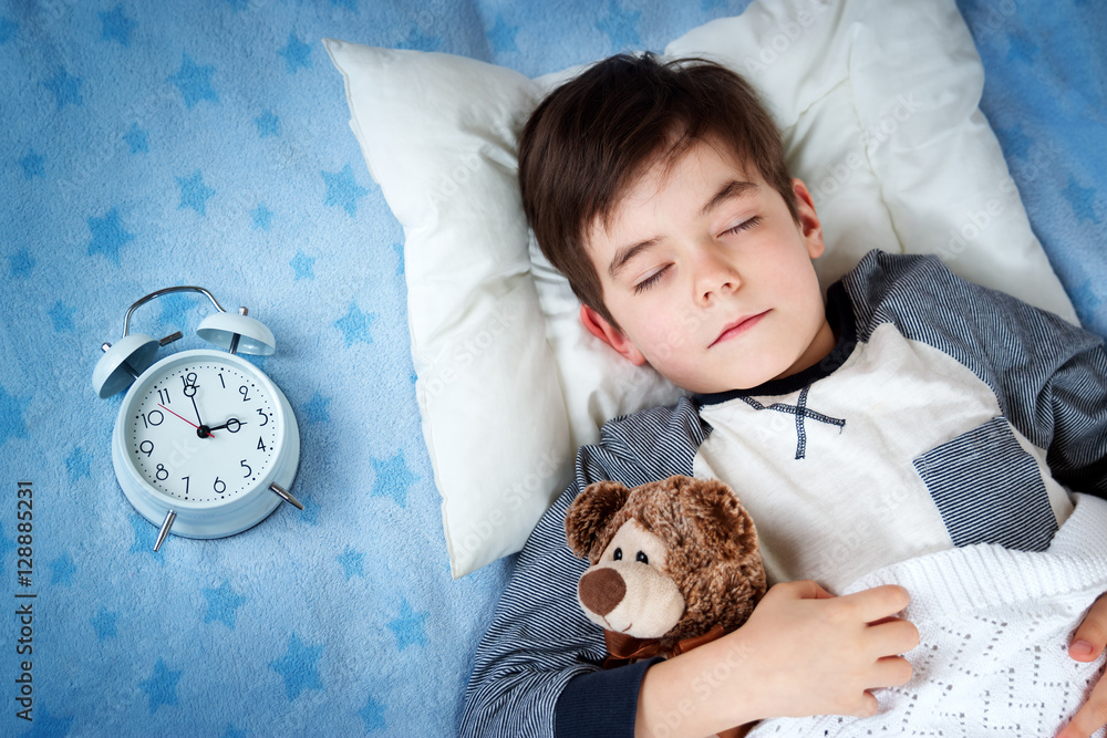 six years old child sleeping in bed on pillow with alarm clock and a teddy bear