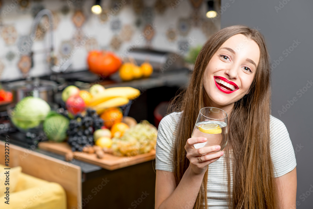 Portrait of a young smiling woman with glass of water in the modern kitchen interior full with fruit