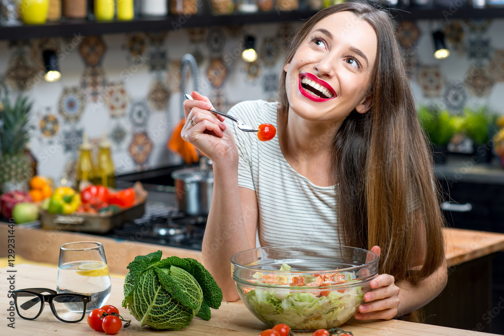 Young smiling woman eating healthy fresh salad in the modern kitchen interior full of fruits and veg