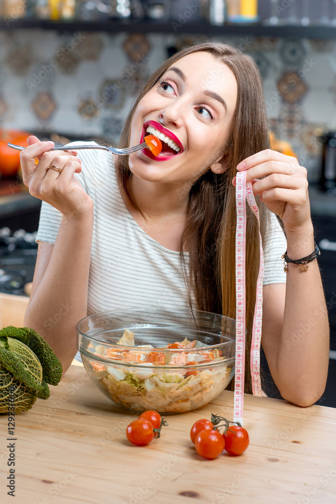 年轻微笑的女人在充满水果和蔬菜的现代厨房里吃着健康的新鲜沙拉