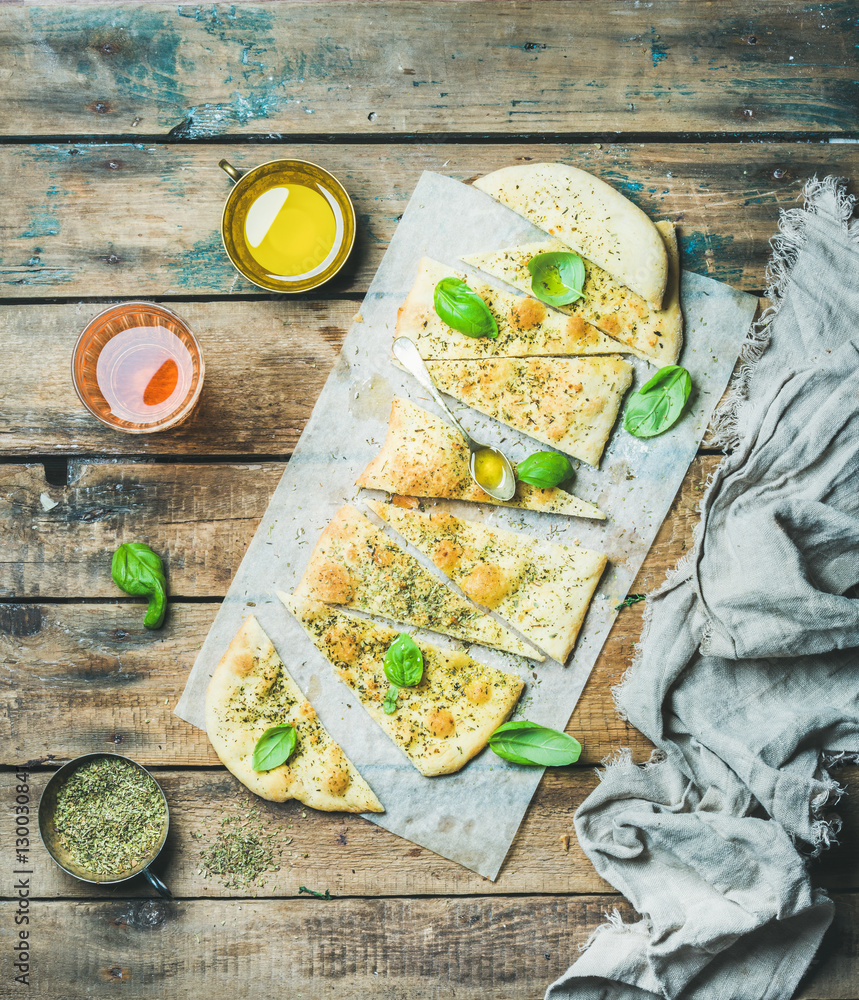 Homemade Italian focaccia flatbread cut into pieces with herbs, fresh basil leaves, olive oil in cup