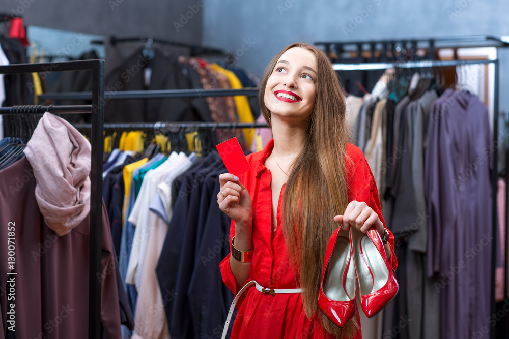 Young happy woman in the red dress holding red shoes and credit card in the luxury clothing store