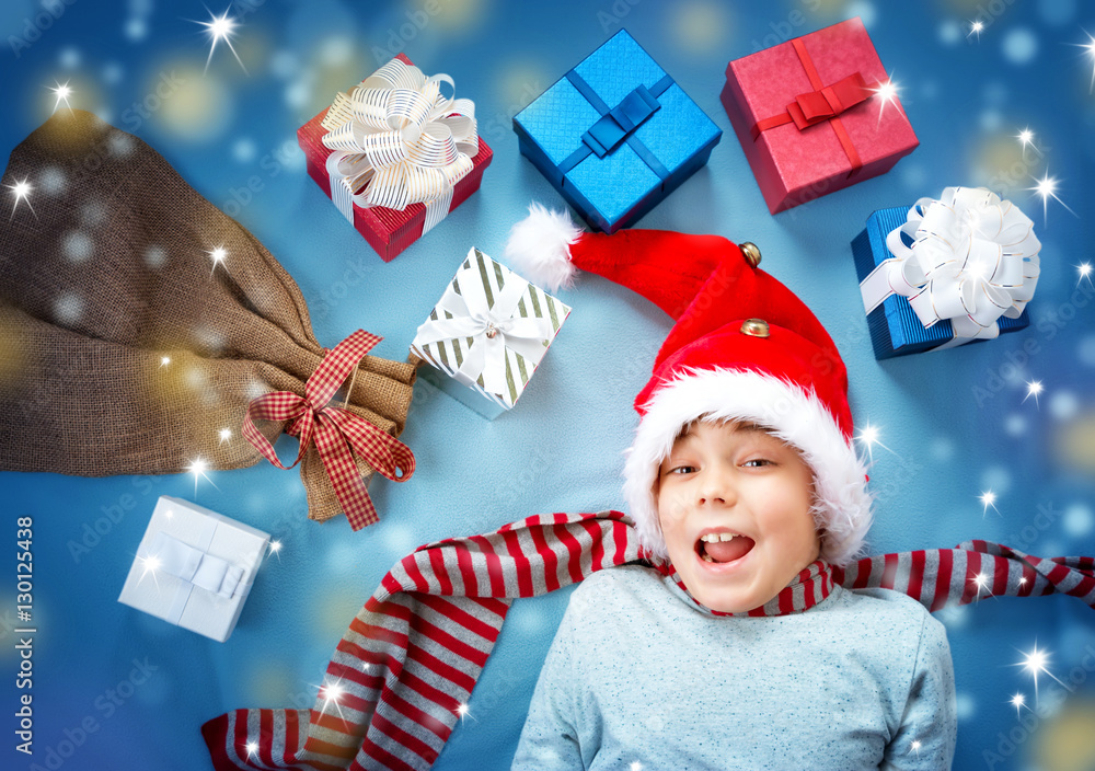 Happy child on blue blanket in red hat holding gift box. Cute boy in christmas cap with many present