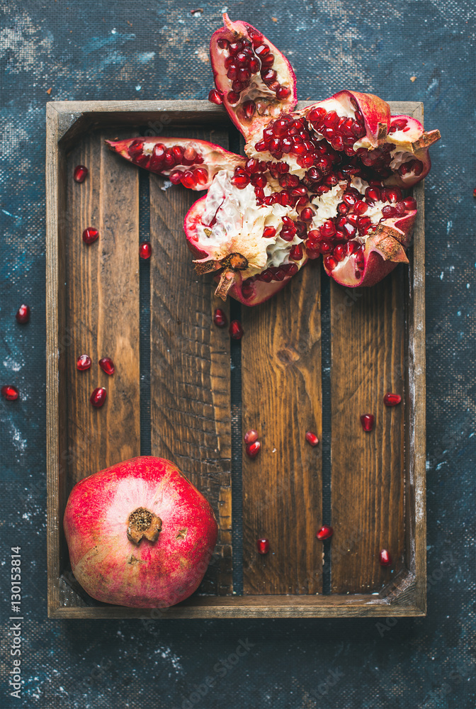 Fresh ripe pomegranates cut in pieces in rustic wooden tray over dark blue plywood background, top v