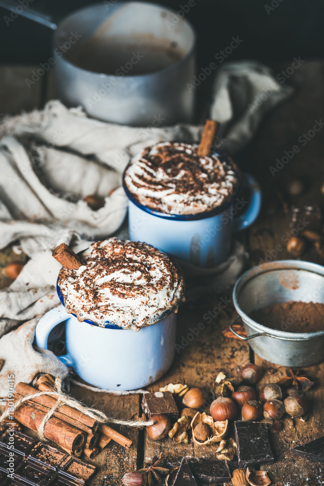 Hot chocolate in blue mugs with whipped cream and cinnamon sticks, spices, nuts and cocoa powder on 