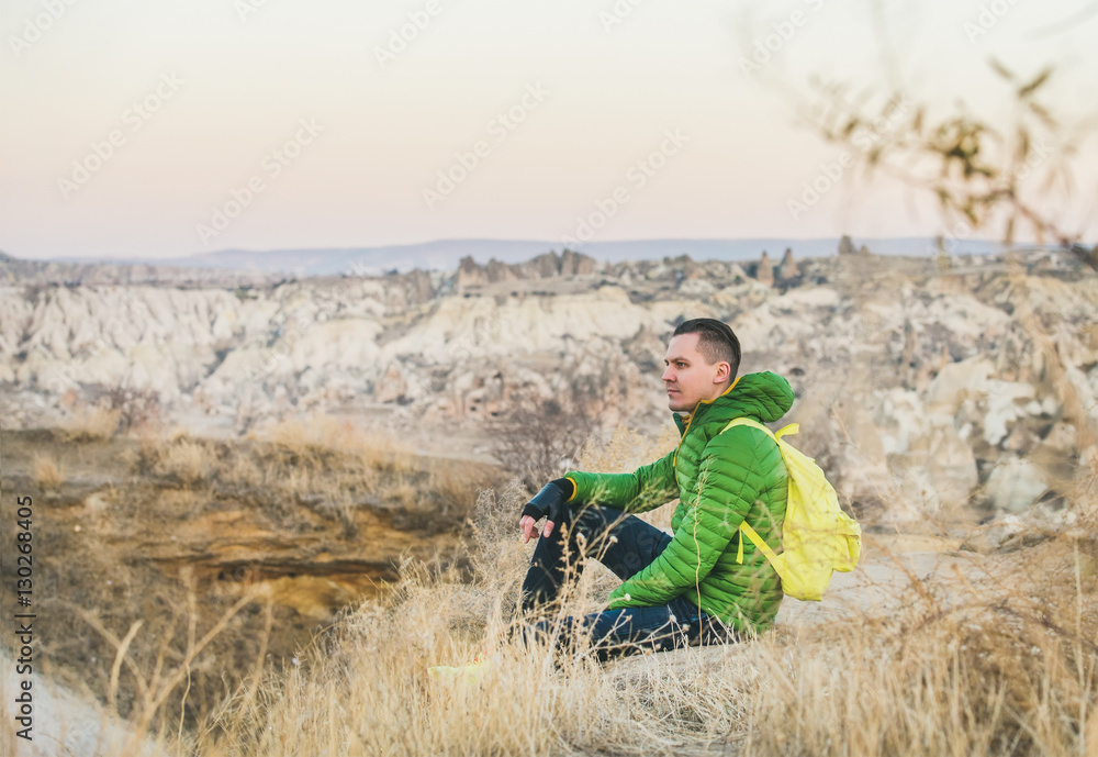 Young man traveller in bright clothes sitting at the edge of cliff in Goreme, Cappadocia, Central Tu