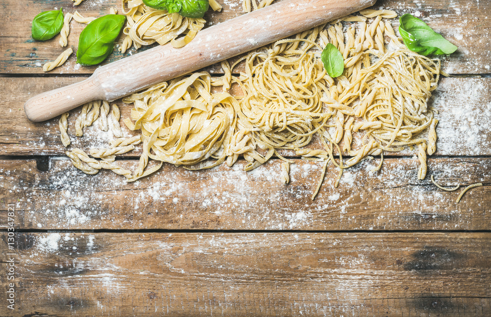 Various homemade fresh uncooked Italian pasta with flour, green basil leaves and plunger on rustic w