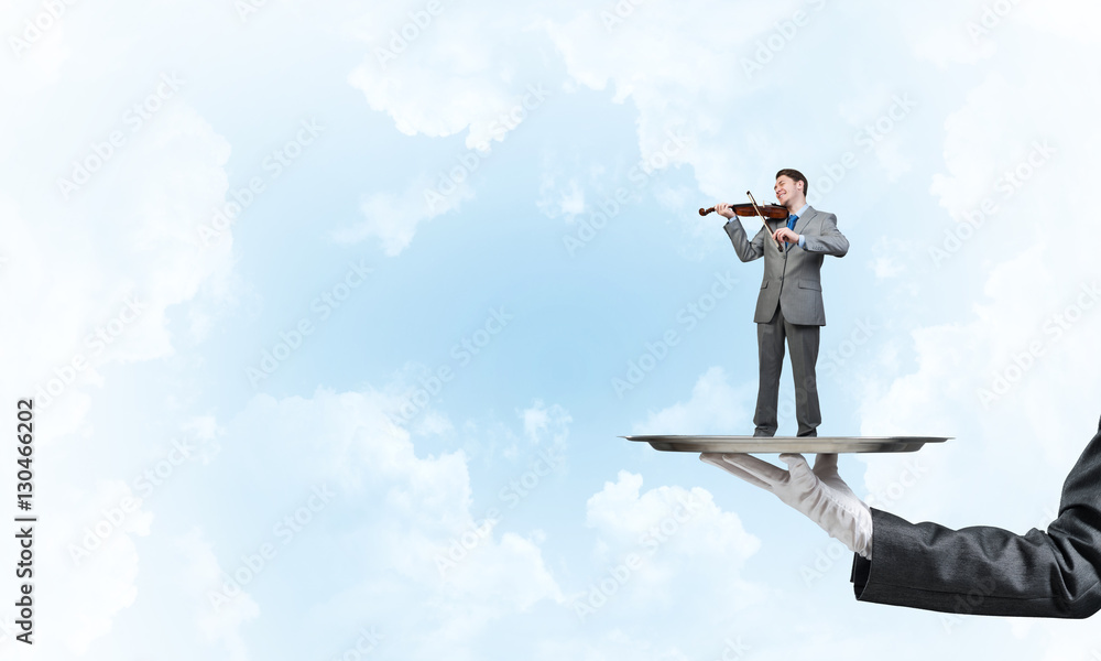 Businessman on metal tray playing violin against blue sky background