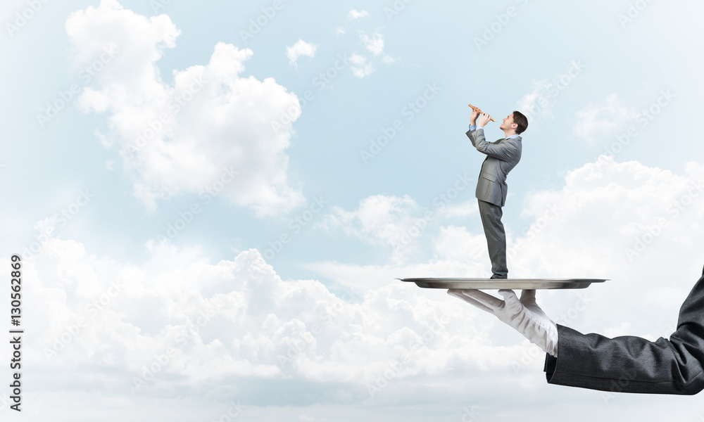 Businessman on metal tray playing fife against blue sky background