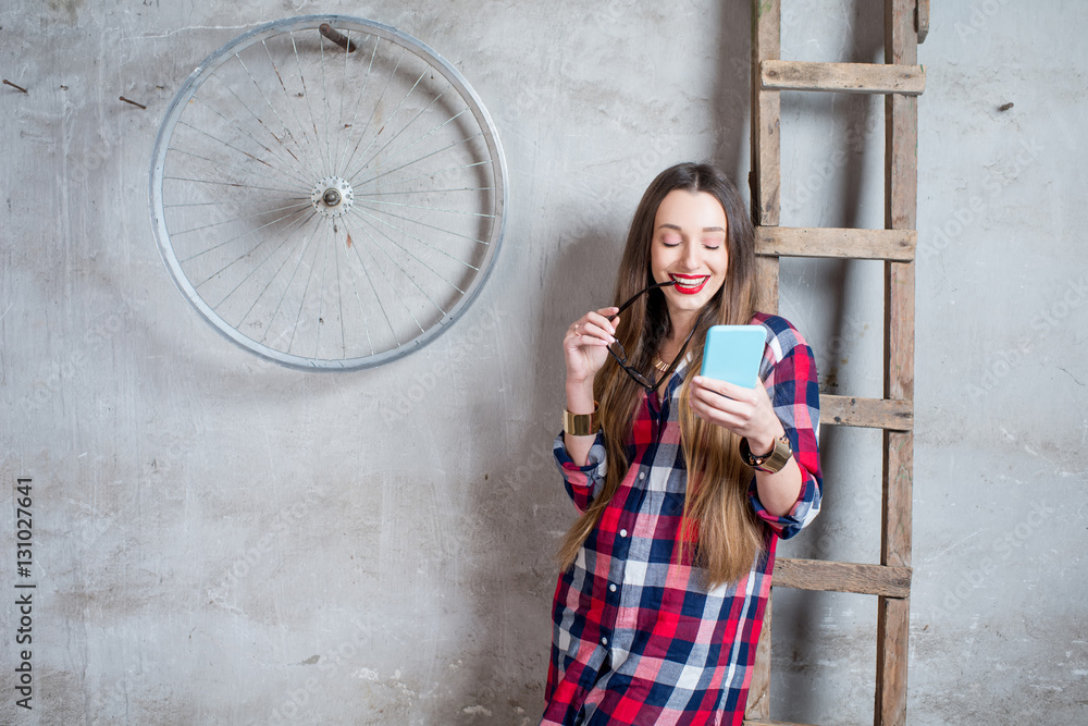 Young woman with phone in checkered shirt standing in the old room with ladder and wheel on the gray