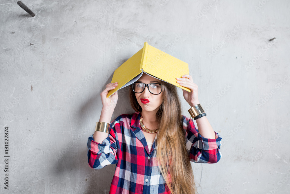 Portrait of confused student in checkered shirt with yellow book on the gray wall background