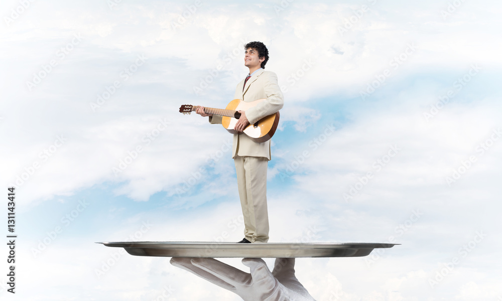 Businessman on metal tray playing acoustic guitar against blue sky background