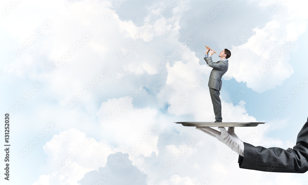 Businessman on metal tray playing fife against blue sky background