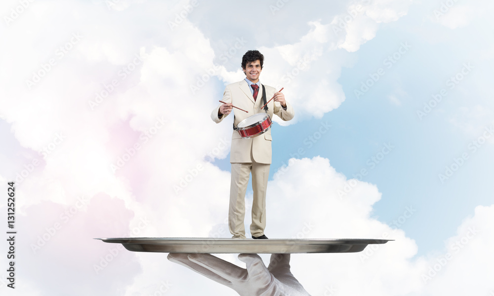 Young businessman on metal tray playing drums against blue sky background