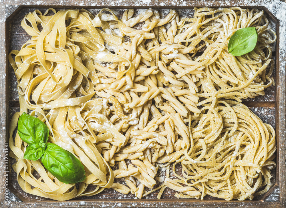 Various homemade fresh uncooked Italian pasta with flour and green basil leaves in wooden tray, top 