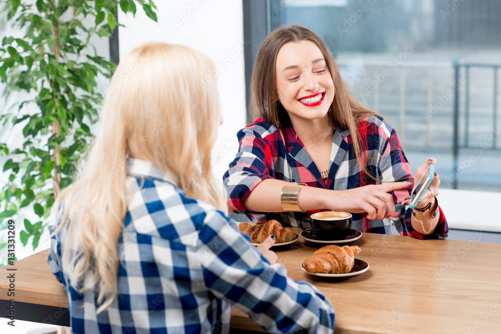 Young female friends dressed casually in checkered shirts sitting with coffee cups and croissants at