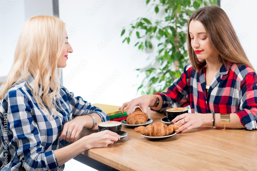 Young female friends dressed casually in checkered shirts sitting with coffee cups and croissants at