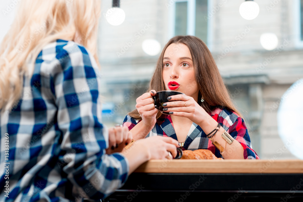 Young female friends dressed casually in checkered shirts sitting with coffee cups and croissants at