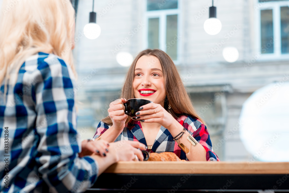 Young female friends dressed casually in checkered shirts sitting with coffee cups and croissants at