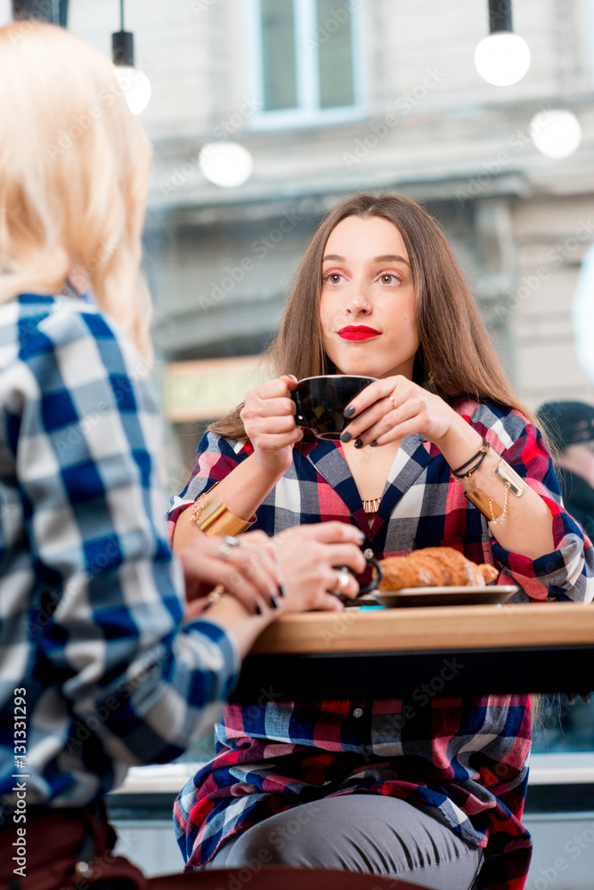 Young female friends dressed casually in checkered shirts sitting with coffee cups and croissants at