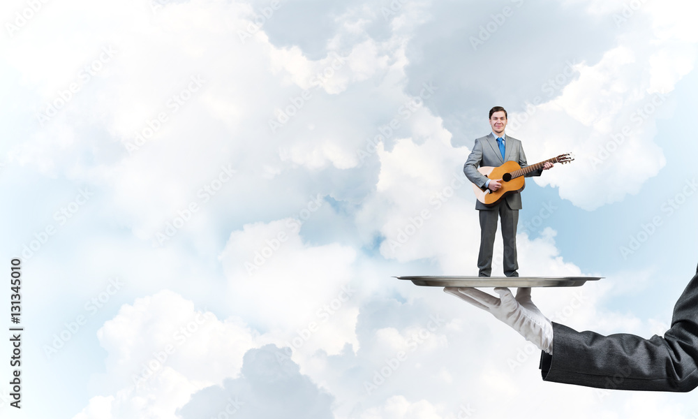 Businessman on metal tray playing acoustic guitar against blue sky background