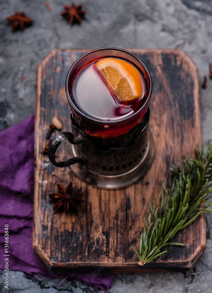 Mulled wine with orange and spices in vintage glass cup holder on wooden board. Concrete background.