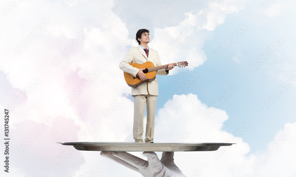 Businessman on metal tray playing acoustic guitar against blue sky background