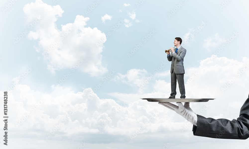 Businessman on metal tray playing fife against blue sky background