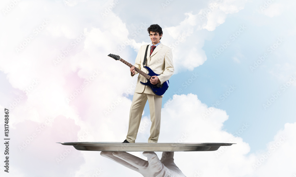 Businessman on metal tray playing electric guitar against blue sky background