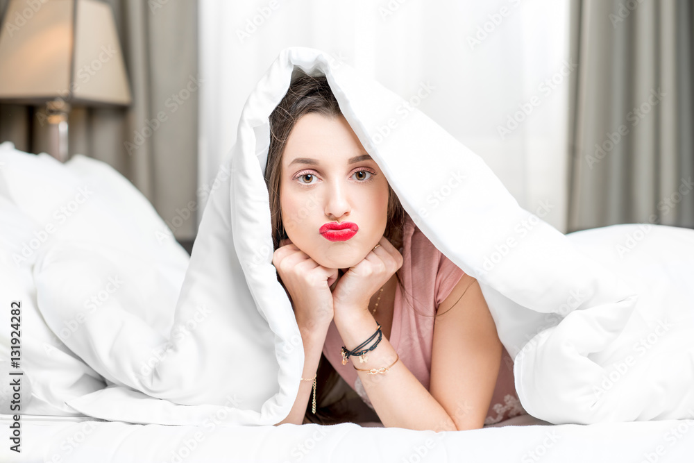 Portrait of young sad woman hidding under the white sheets on the bed