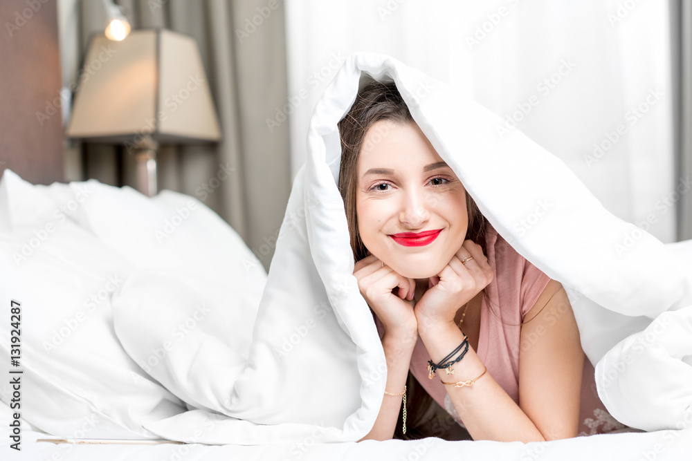 Portrait of young smiling woman hidding under the white sheets on the bed