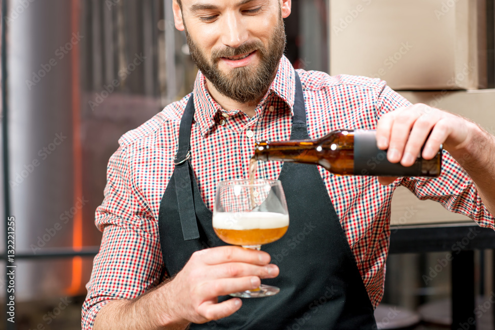 Handsome brewer in apron and shirt pouring beer into the glass at the manufacturing. Image focused o