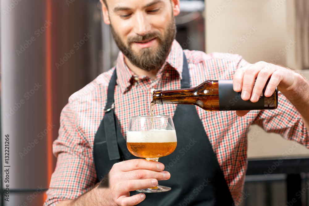 Handsome brewer in apron and shirt pouring beer into the glass at the manufacturing. Image focused o