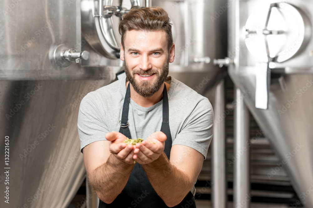 Portrait of a handsome brewer in uniform smelling hop at the manufacturing with metal tanks on the b