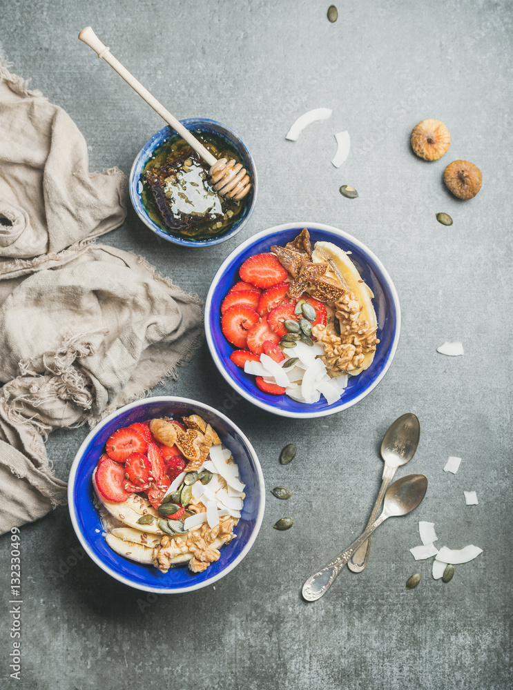 Yogurt, granola, seeds, fresh and dry fruits and honey in blue ceramic bowls over grey concrete back