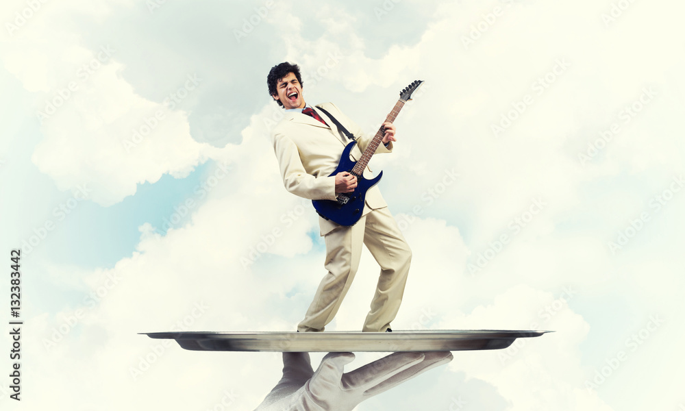 Businessman on metal tray playing electric guitar against blue sky background