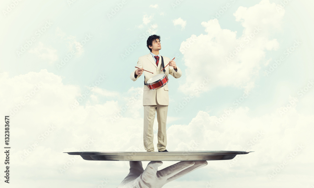 Young businessman on metal tray playing drums against blue sky background