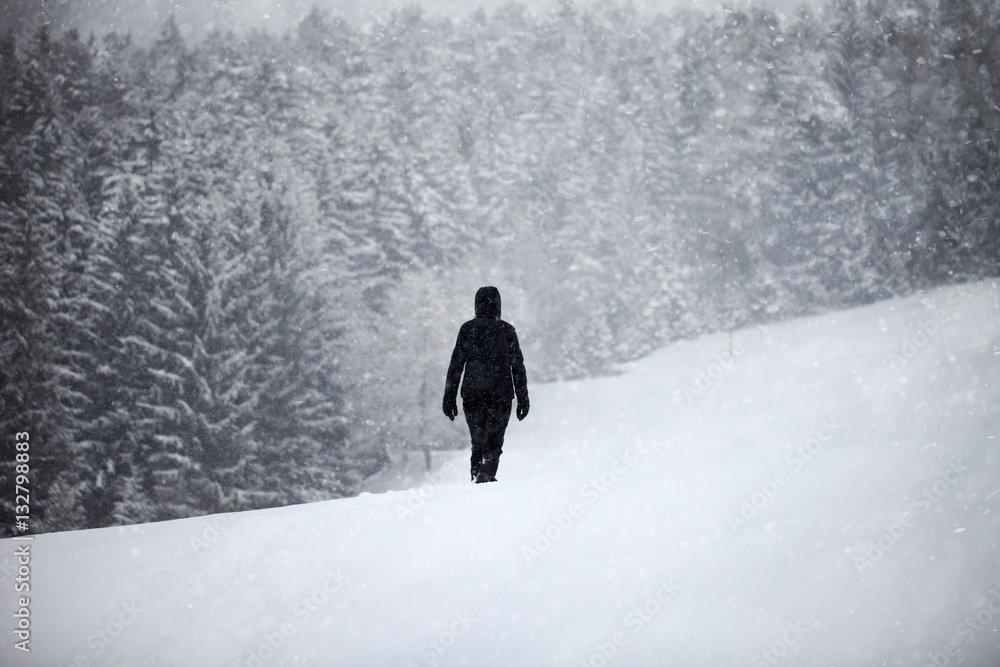 Woman in black dress walking alone across the snowy landscape during snowfall.