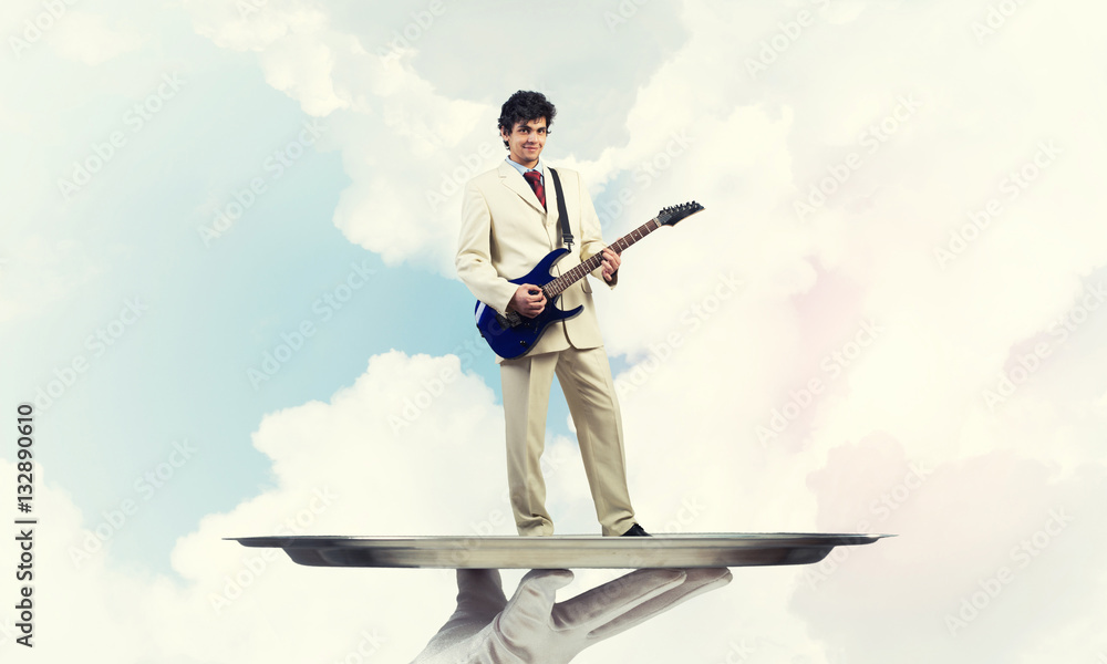 Businessman on metal tray playing electric guitar against blue sky background