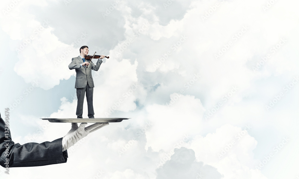 Businessman on metal tray playing violin against blue sky background