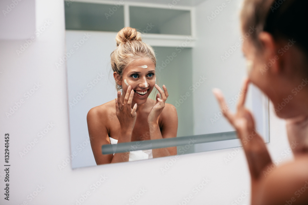 Happy young woman applying cream in bathroom