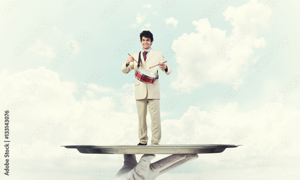 Young businessman on metal tray playing drums against blue sky background