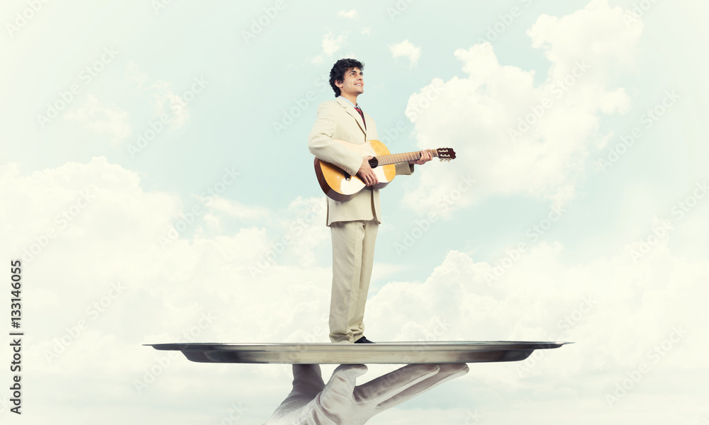 Businessman on metal tray playing acoustic guitar against blue sky background