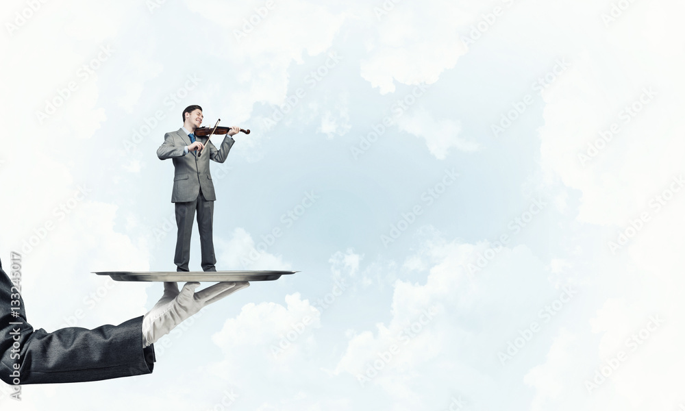 Businessman on metal tray playing violin against blue sky background