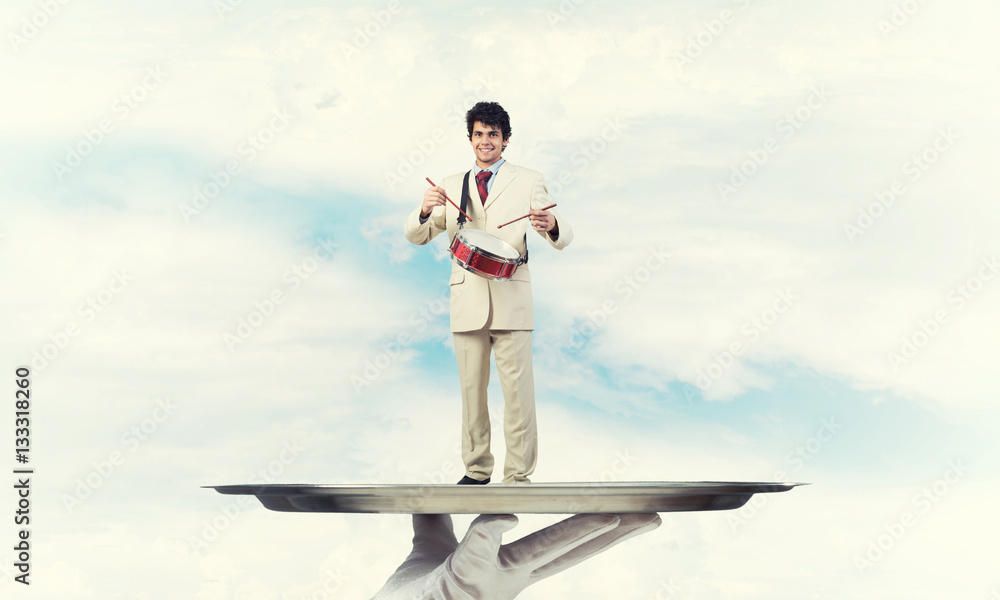 Young businessman on metal tray playing drums against blue sky background