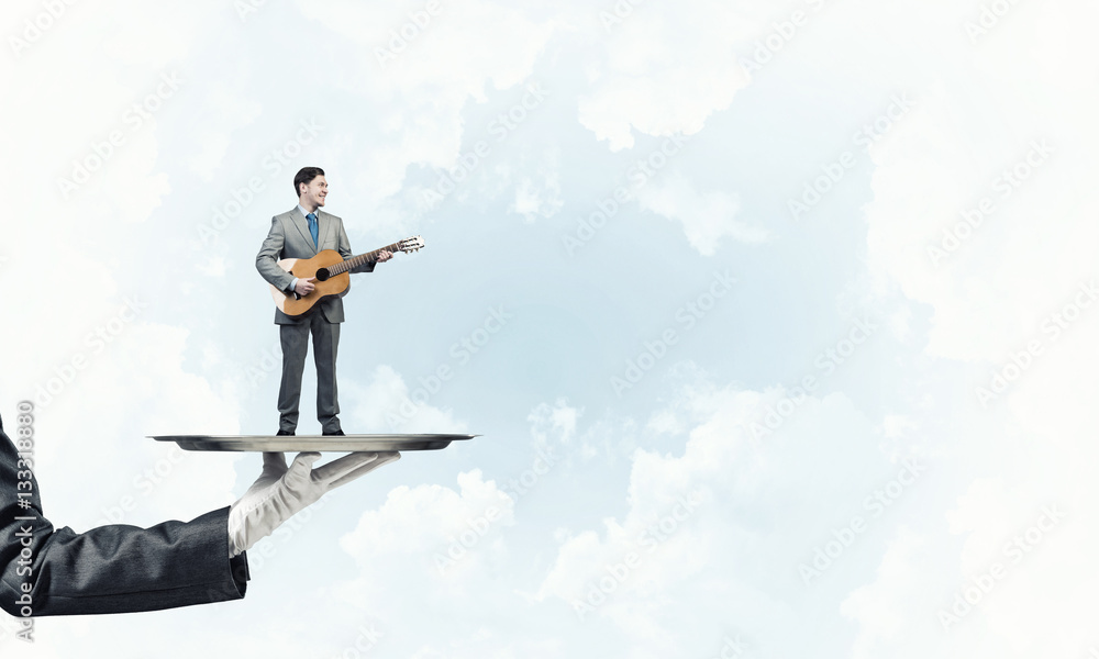 Businessman on metal tray playing acoustic guitar against blue sky background