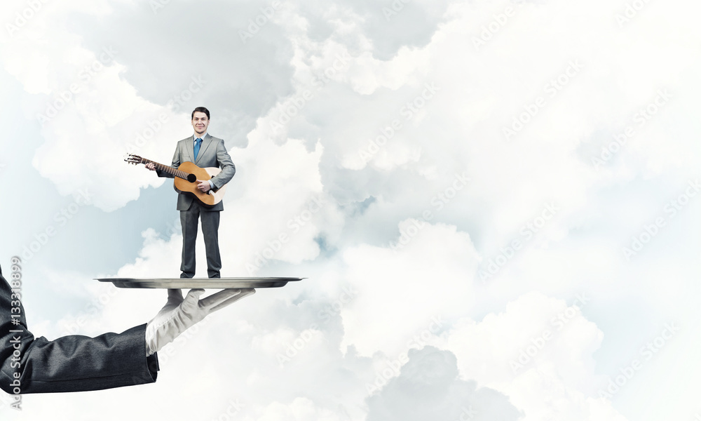 Businessman on metal tray playing acoustic guitar against blue sky background