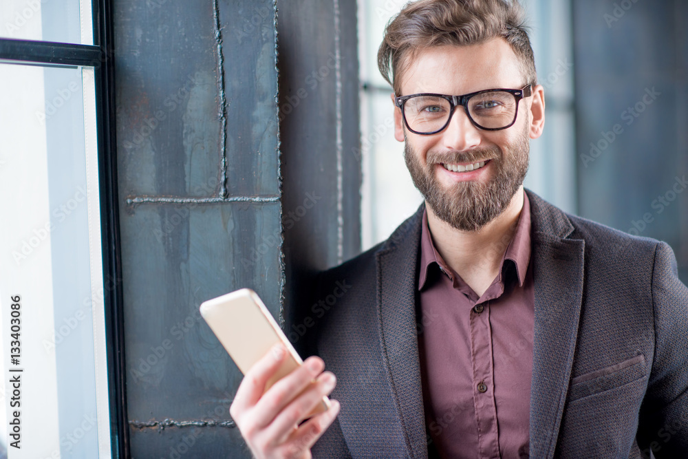 Portrait of caucasian bearded businessman dressed in suit with eyeglasses holding smart phone near t