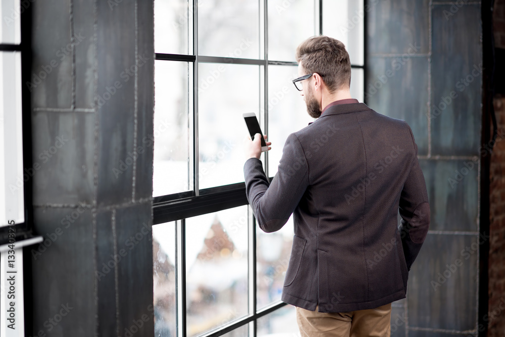 Businessman dressed in the suit standing with smart phone near the big window in the loft interior s